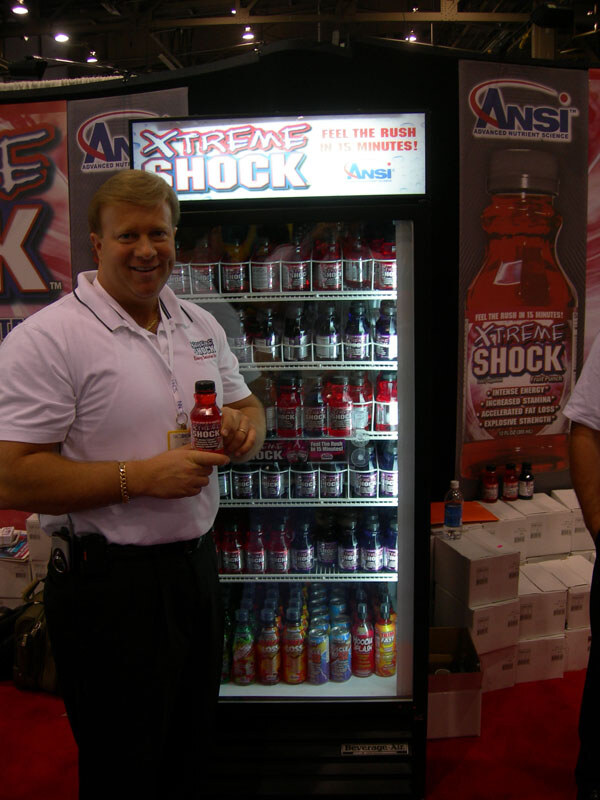 A smiling man in a white polo holds a bottle of Xtreme Shock beside a lit refrigerator filled with bottles, with ANSI banners behind him.