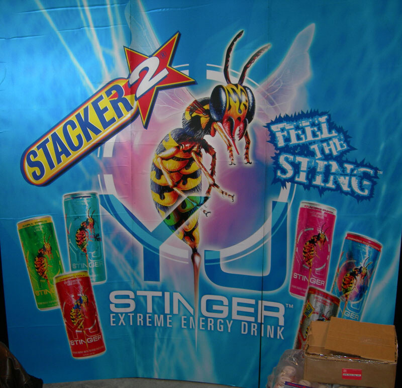 A blue banner shows a stylized wasp, the Stacker 2 logo, the words Feel the Sting and YJ Stinger Extreme Energy Drink, with colorful cans around it; cardboard boxes sit at lower right.
