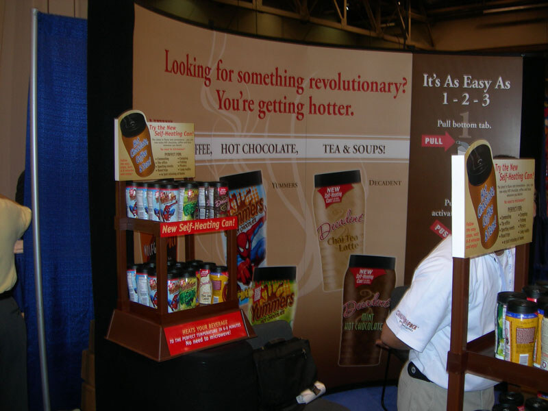 A booth backdrop reads Looking for something revolutionary? You're getting hotter, with Yummers and Decadent self-heating cans on display shelves and a person in a white shirt at right.