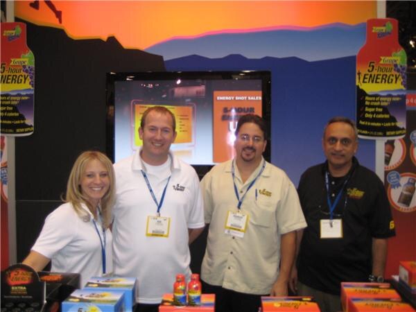 A woman and three men wearing lanyards stand together at a booth with 5-hour Energy bottle graphics, a screen and product boxes and bottles in front of them.