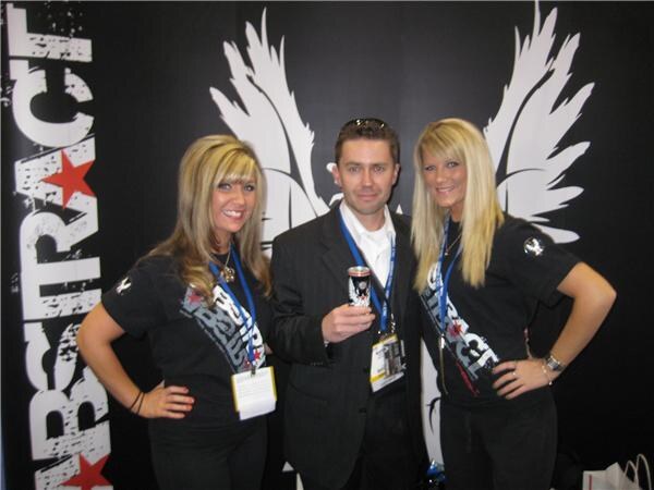 A man in a dark suit holds an energy drink can between two smiling women in black Abstract shirts, all wearing lanyards, in front of a black backdrop with white wings and Abstract lettering.