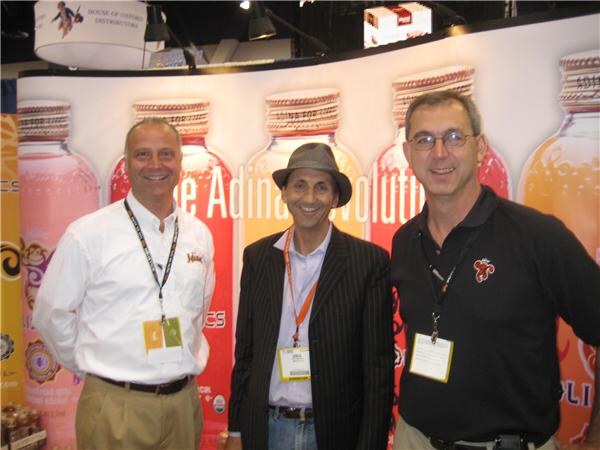 Three smiling men with lanyards stand in front of a display of large bottle graphics and Adina signage. The middle man wears a gray fedora and pinstriped jacket.