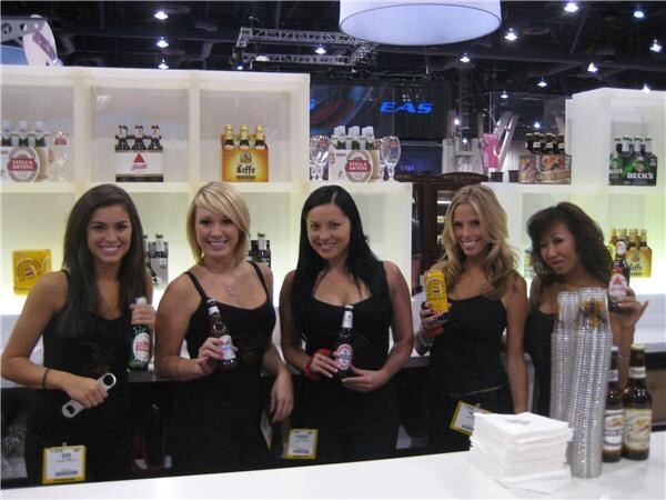 Five smiling women in black tops stand behind a white bar holding beer bottles, with shelves of Stella Artois and Beck's bottles behind them and stacks of cups and napkins on the counter.