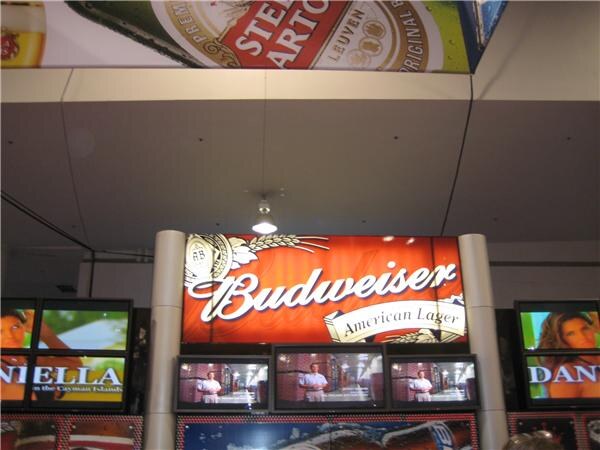 Booth display with a lit red Budweiser American Lager sign above three video screens, flanked by more screens, and a Stella Artois banner hanging overhead.