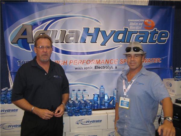 Two men stand in front of an AquaHydrate banner and cases of bottled water; one wears glasses and a black shirt, the other a hat with sunglasses, a light blue shirt and a badge.