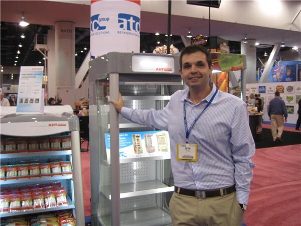 A smiling man in a light checked shirt and lanyard stands with a hand on an open-front AHT refrigerated cabinet, with an ATC refrigeration sign overhead and a snack-stocked unit at left.