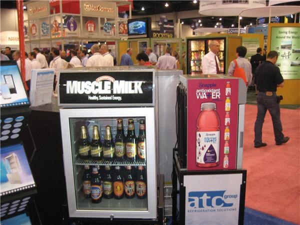 A small glass-door cooler topped with a Muscle Milk sign holds bottles, beside a Snapple antioxidant water display and an atc group sign, with attendees in the aisle behind.