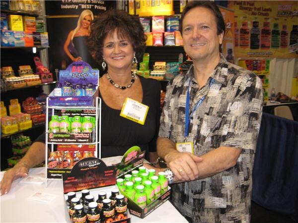 A woman with dark curly hair and a man in a patterned shirt stand behind a table with a Bad Energy shot display and other energy shot products, with shelves of products behind them.
