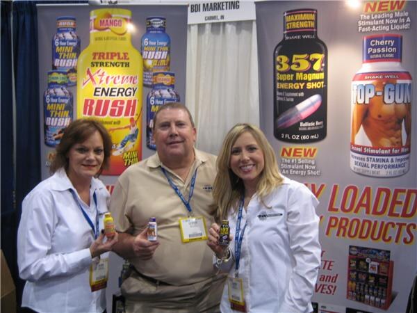 Two women and a man stand at the BDI Marketing booth holding small energy shots, in front of banners for Xtreme Energy Rush, Mini Thin Rush, 357 Super Magnum and Top-Gun.