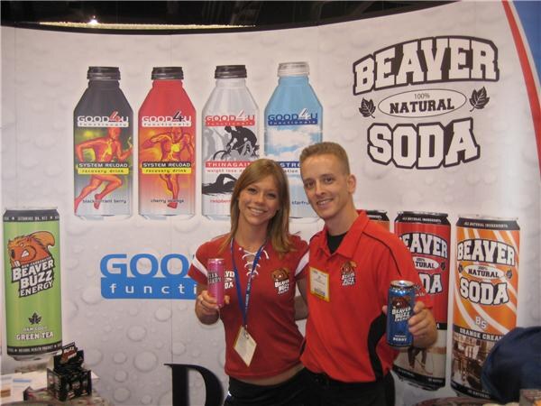 A woman and a man in red Beaver Buzz Energy shirts hold cans in front of a backdrop of Beaver Soda, Beaver Buzz Energy and Good 4 U Functionals bottles and cans.