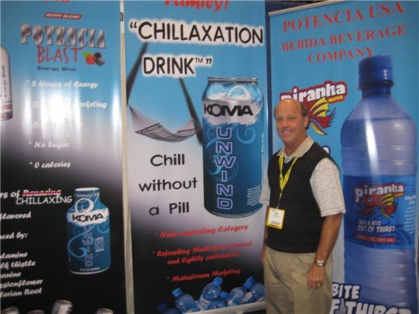 A smiling man with a lanyard stands beside banners for Potencia Blast, Koma Unwind and Piranha Water, with Potencia USA and Bebida Beverage Company text at the right.