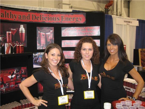 Three smiling women in black Bing shirts, two with badges, stand at a booth with red and black signs about energy drink, with cans stacked in front and cups on a tray.