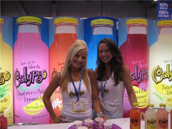 Two smiling women in white tops and lanyards stand behind a counter with small cups, in front of large Calypso lemonade bottle graphics including Raspberry Pink Lemonade.