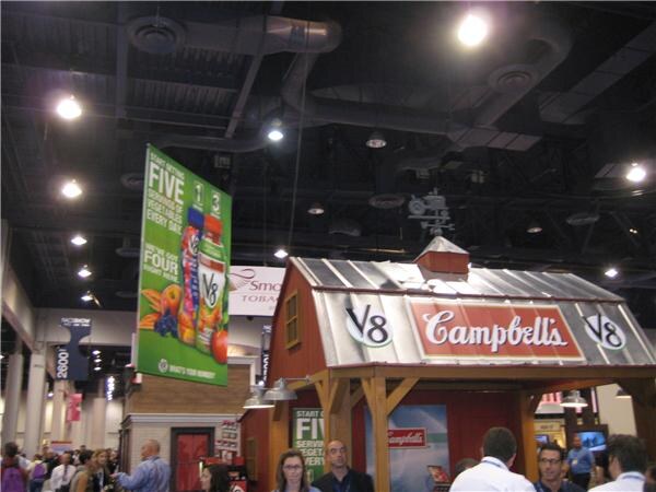 A barn-shaped booth with Campbell's and V8 signs stands under a high ceiling in an exhibit hall, with a hanging V8 banner and attendees below.