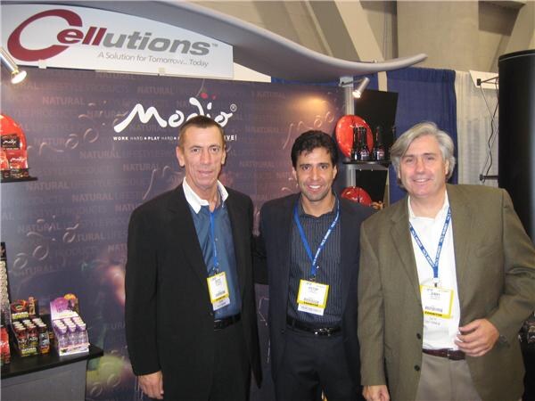 Three men in jackets and badges stand in front of a Cellutions booth with a Modjo backdrop, with product displays on both sides.
