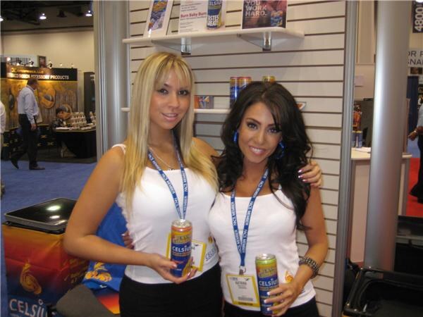 Two women in white tops and lanyards each hold a can of Celsius in front of a shelved booth wall displaying more cans.