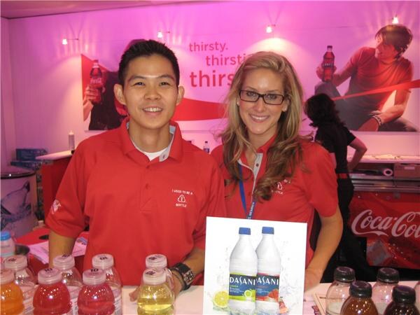 A man and a woman in red polo shirts stand behind a counter of bottled drinks and a Dasani flavored water sign, with Coca-Cola graphics behind them.