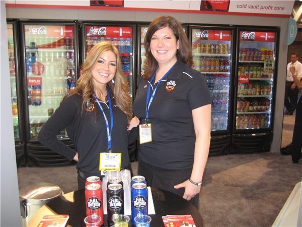 Two women in dark tops and lanyards stand at a table with Full Throttle cans, in front of Coca-Cola coolers stocked with beverages.