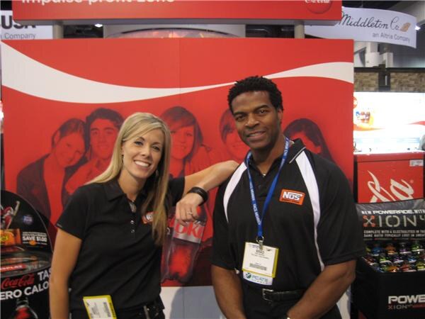 A woman in a black polo and a man in a black NOS polo with a lanyard stand before a red Coca-Cola display, with Powerade signage at right.
