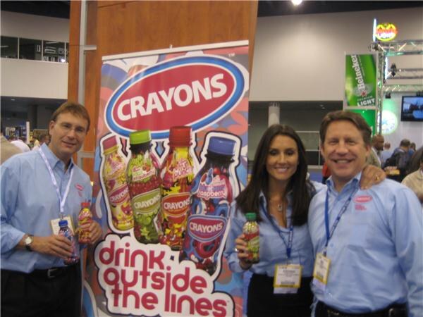 Three people in light blue shirts and lanyards pose in front of a Crayons banner reading "drink outside the lines", holding Crayons bottles. A Heineken Light sign is in the background.