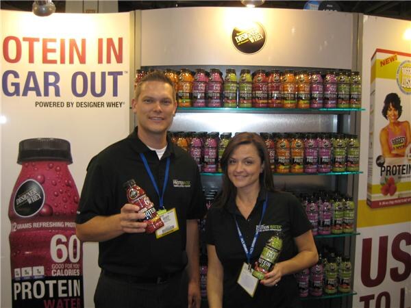 A man and a woman in black shirts and lanyards each hold a bottle of protein water in front of shelves of Designer Whey bottles and a banner reading "Protein in, gar out".