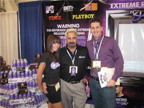 A woman in a black top and two men, one in a black shirt and one in a purple shirt, pose in a booth with stacked purple cans and a backdrop showing MTV, BET Networks, Playboy and Time logos.