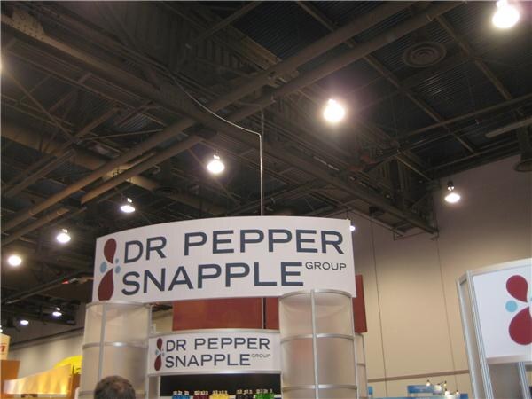 A curved white sign reading Dr Pepper Snapple Group hangs above a booth under an exposed ceiling with lights. A smaller sign with the same name is on the booth below.