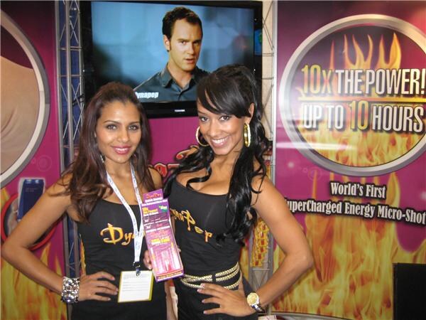 Two women in black Dynapep tops pose at a booth with a flame graphic reading "10x the power! Up to 10 hours". One holds a small Dynapep package, and a TV is behind them.