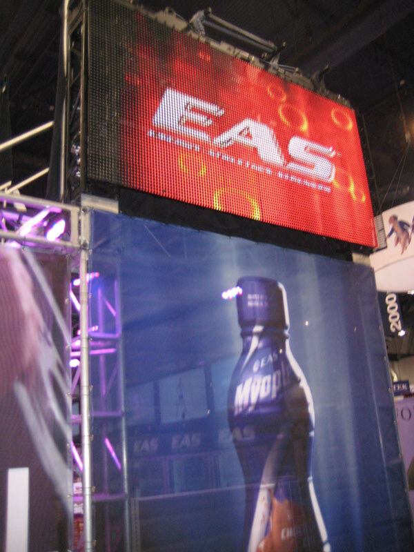 A tall LED screen displaying the EAS logo on a red background above a large banner of a Myoplex bottle, mounted on a truss at a trade show booth.