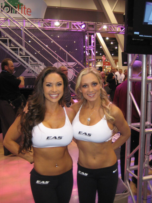 Two smiling women in white EAS sports tops and black EAS pants stand side by side in front of a truss and staircase, with trade show attendees visible behind them.