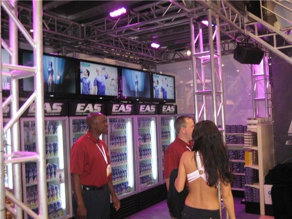 A man in a red shirt and lanyard and a second man in a red shirt stand beside a woman with long dark hair, facing a row of EAS-branded coolers stocked with drinks, with video screens above.