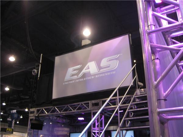 A large projection screen showing the EAS logo and the words Energy, Athletics, Strength, mounted above a metal truss structure with a staircase railing in a trade show hall.