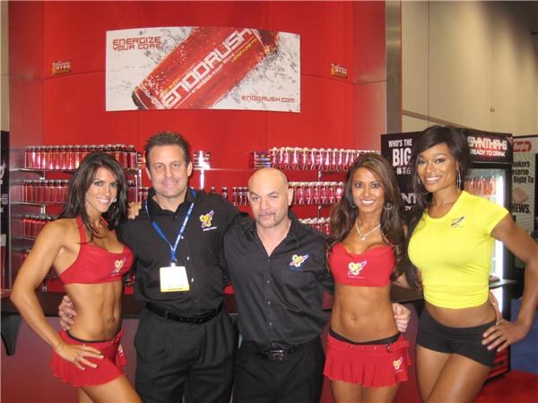 Two men in black shirts stand with three women in red or yellow outfits in front of a red display of EndoRush cans and a banner reading EndoRush.