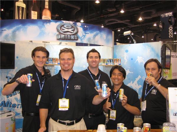 Five men in black Ex Drinks shirts and lanyards stand together at a booth, several holding cans of the drink, in front of a blue Ex Drinks backdrop.