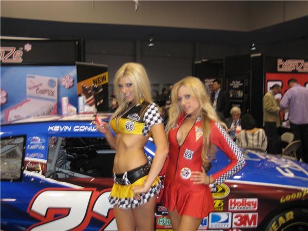 Two women in checkered racing-style outfits, one in yellow and one in red, stand beside a blue race car with Holley, 3M, DirecTV and Goodyear logos near an Extenze display.