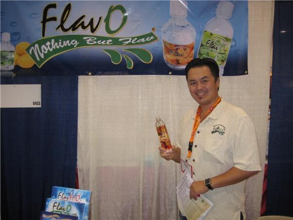 A smiling man with an orange lanyard holds a bottle of FlavO water in front of a FlavO banner reading "Nothing But Flav", with product brochures in the foreground.