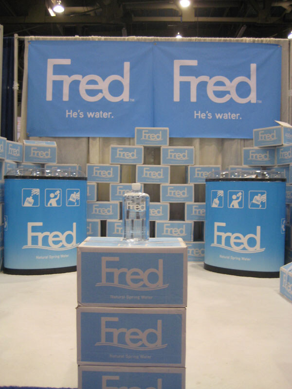 Fred booth with two blue banners reading "Fred He's water.", stacked blue Fred cartons, two blue round displays and a bottle of water on top of cases of Fred Natural Spring Water.