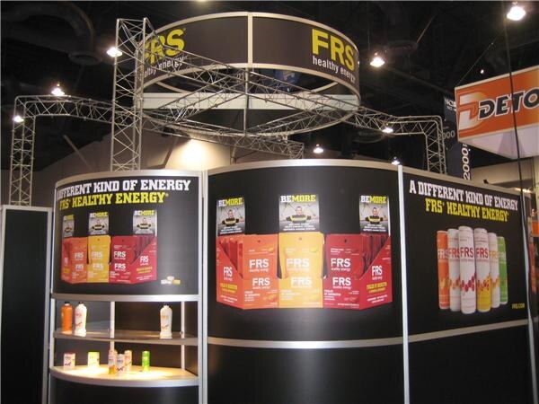 FRS booth with a circular overhead sign, black display walls reading "A different kind of energy," and shelves of FRS healthy energy chews, packets and bottles in orange and red packaging.