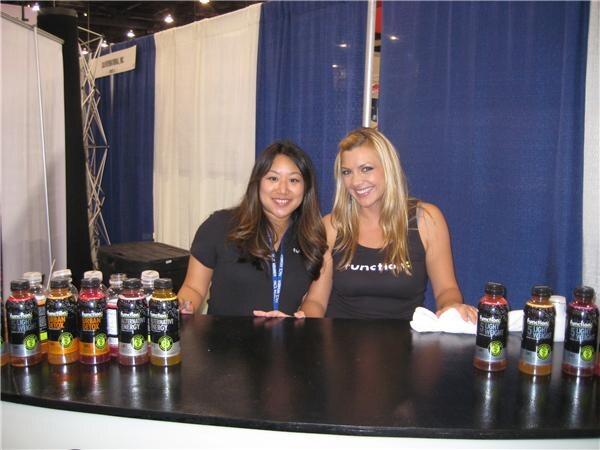 Two smiling women in black tops stand behind a black counter lined with bottles of Function drinks in several flavors, with blue and white curtains behind them.