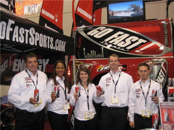 Five people in white shirts and lanyards hold red Go Fast energy drink cans in front of a red Go Fast truck and a black GoFastSports.com canopy.