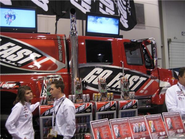 A red Go Fast! truck with the brand logo sits behind displays of Go Fast! energy drink cans and price signs, while a young man and woman in white shirts talk in front of it.