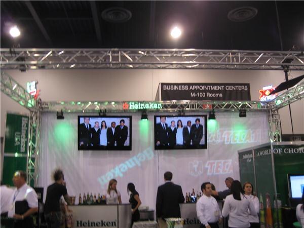 Heineken and Tecate booth under a truss, with two screens showing groups of people in dark clothing, a Business Appointment Centers sign, and people standing near the counter.