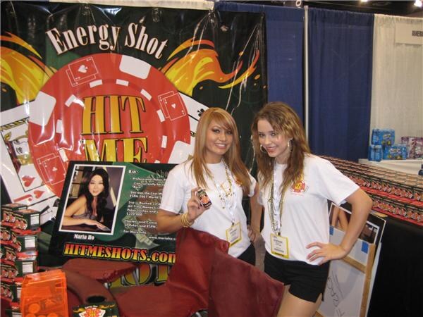 Two young women in white T-shirts and badges stand at the Hit Me Energy Shot booth; one holds a small bottle. A poker chip banner and HITMESHOTS.com sign are behind them.