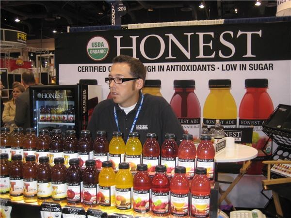 A man with glasses and a lanyard stands behind rows of Honest Ade bottles in several colors, in front of a large Honest banner and a small Honest-branded refrigerator.