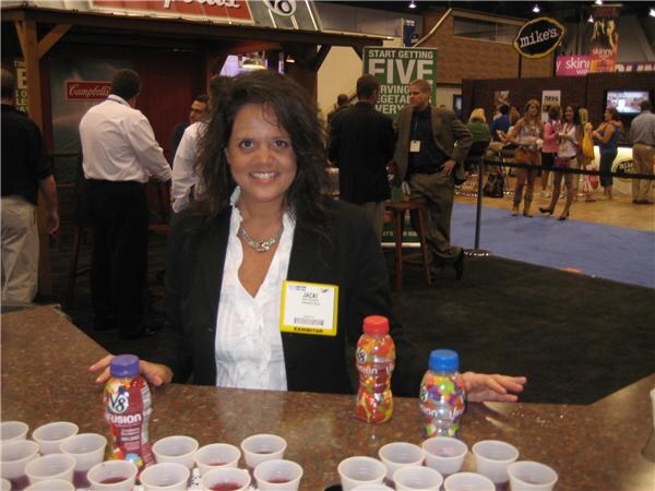 Jacki, wearing an exhibitor badge, smiles behind a counter with V8 Fusion bottles and rows of small sample cups. Other attendees stand in the background.