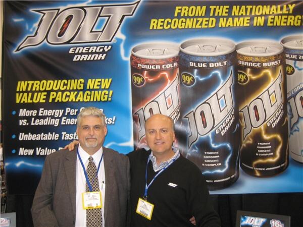 Two men in lanyards stand side by side in front of a Jolt Energy Drink banner showing cans in several flavors and the text "Introducing new value packaging!"