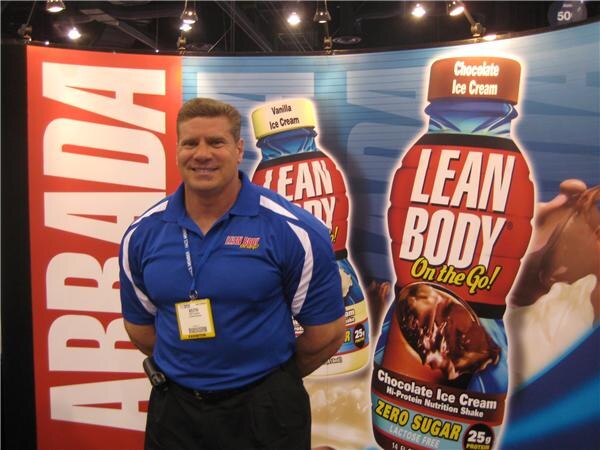 A smiling man in a blue polo with a badge stands in front of large Lean Body "On the Go!" chocolate and vanilla ice cream shake bottle graphics and a red banner.