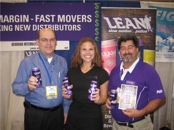 Two men and a smiling woman hold purple Lean cans in a booth with a Lean banner and a purple sign reading "Margin - Fast Movers"; the man on the right holds a 2010 brochure.