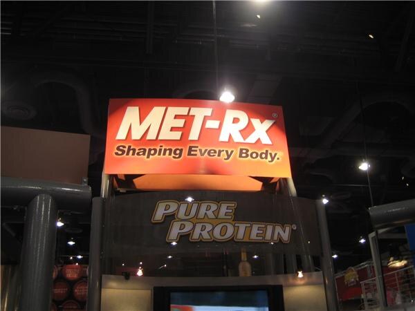 An orange MET-Rx sign reading "Shaping Every Body." hangs above a Pure Protein sign on a booth structure under a dark convention hall ceiling.