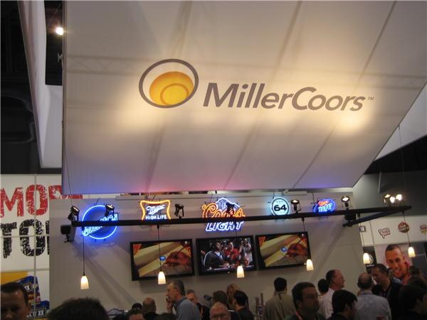 A MillerCoors logo sign hangs above a bar with neon signs for Miller High Life and Coors Light, three television screens and a crowd of attendees below.